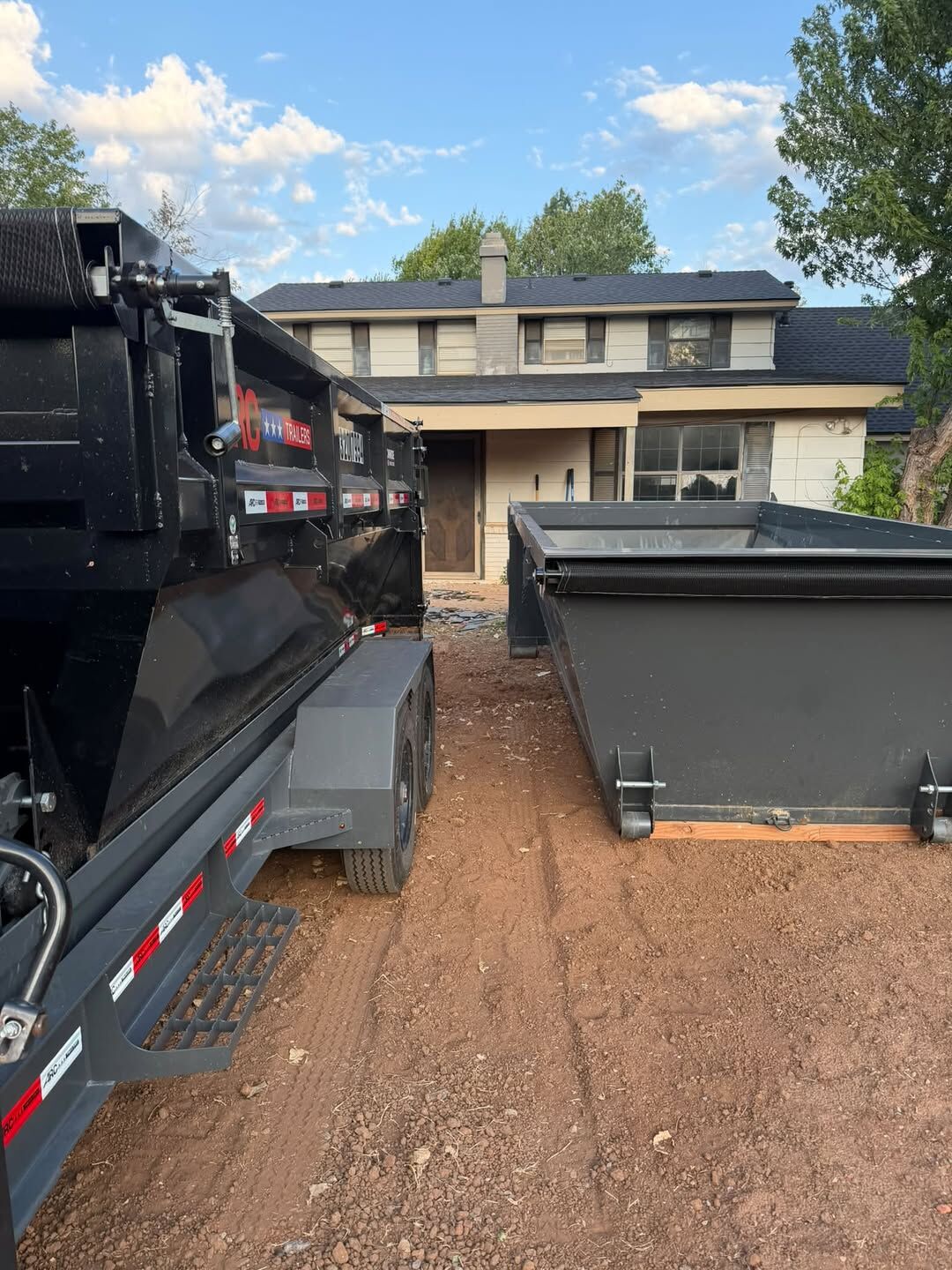 Two SCORO roll-off bins on red dirt in a driveway, one loaded on the trailer and one set on the ground
