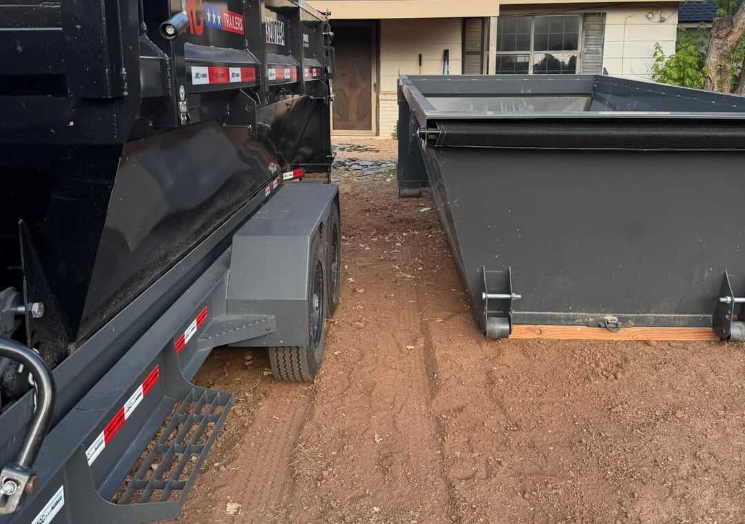 Two SCORO roll-off bins on red dirt in a driveway, one loaded on the trailer and one set on the ground